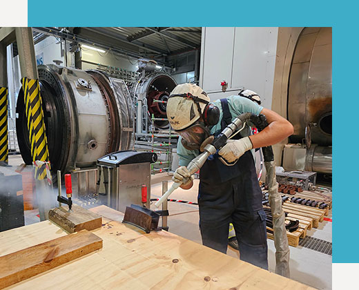 Pracownik Pamech ubrany w odzież ochronną używa dużego narzędzia do piaskowania lub czyszczenia metalowego elementu na stole warsztatowym w obiekcie przemysłowym z ciężkimi maszynami w tle.