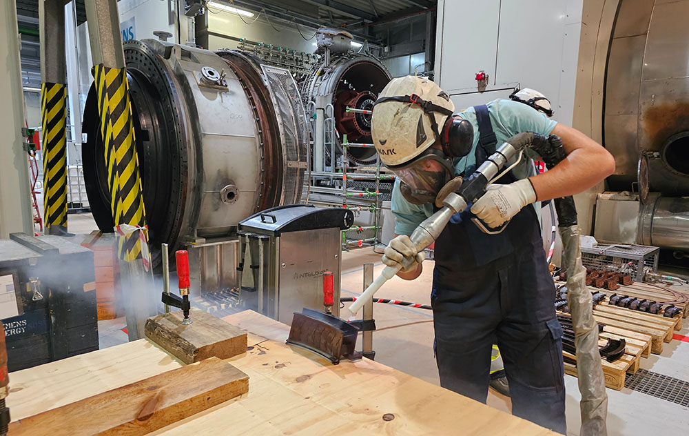 Pracownik w odzieży ochronnej i kasku używa dużego narzędzia przypominającego wąż do realizacji czyszczenia suchym lodem na metalowej części umieszczonej na drewnianym stole w warsztacie przemysłowym z dużymi maszynami w tle.