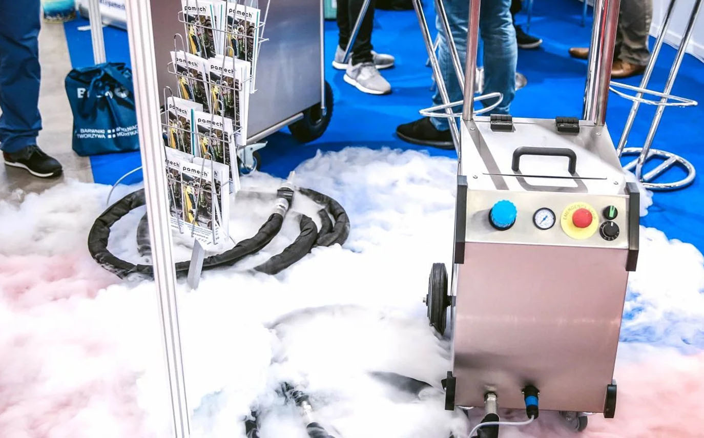 Maszyna do mgły emituje gęstą białą parę na niebieską podłogę wyłożoną dywanem podczas imprezy w pomieszczeniu, widoczne są przyciski sterujące i wskaźniki; w tle stoją ludzie. Idealne rozwiązanie dla obiektów oferujących sprzedaż granulatu suchego lodu w celu zwiększenia efektów.