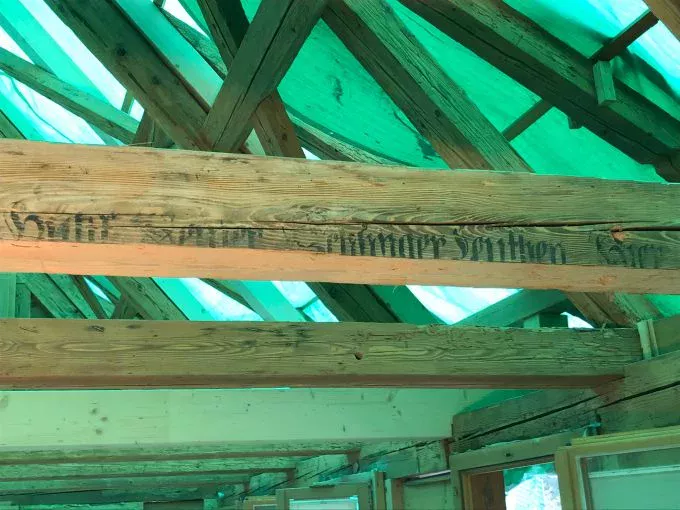 Odsłonięte drewniane belki w budowanym dachu, z wyblakłym czarnym tekstem na jednej belce. Dach przykryty jest zieloną plandeką, przez którą wpada naturalne światło - idealne warunki do czyszczenie elewacji suchym lodem.