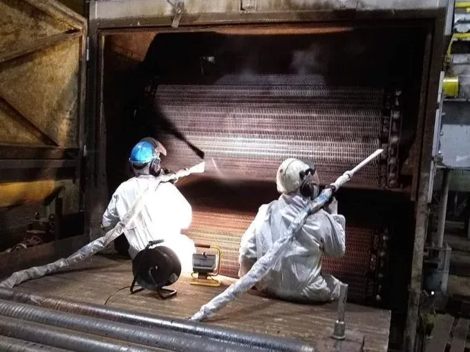 Dwóch pracowników w kombinezonach ochronnych i kaskach używa węży do czyszczenia dużych urządzeń przemysłowych wewnątrz obiektu. Para lub mgła unosi się podczas wykonywania usługi czyszczenia suchym lodem, a sprzęt ochronny jest widoczny przez cały czas.