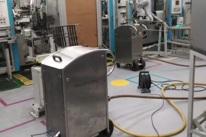 Pomieszczenie czyste ze sprzętem przemysłowym, maszynami i kablami na podłodze; osoba w białym kombinezonie ochronnym pracuje w tle wśród urządzeń ze stali nierdzewnej i maszyn w niebieskim kolorze, ilustrując schematyczne działanie zaawansowanej produkcji.