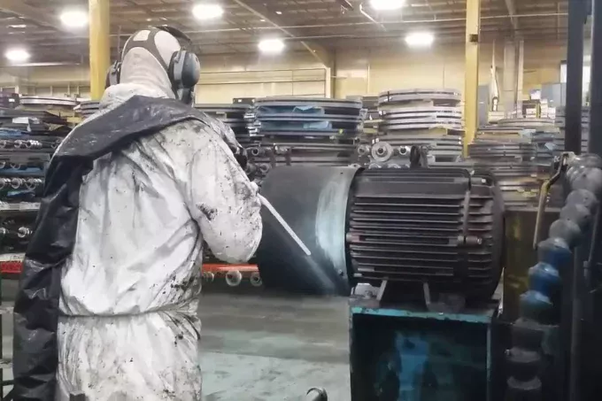 Osoba w odzieży ochronnej i słuchawkach używa urządzenia natryskowego do usuwania pleśni suchym lodem na dużej maszynie przemysłowej wewnątrz fabryki z metalowymi półkami i sprzętem w tle.