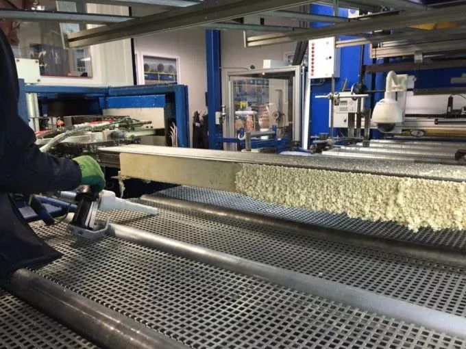 Pracownik w zielonych rękawicach używa narzędzia do obsługi dużego metalowego panelu pokrytego pianką izolacyjną, podkreślając Utrzymanie Ruchu i Konserwację w otoczeniu przemysłowym z metalowymi kratami i niebieskimi maszynami.